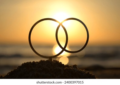 Black outlines of wedding rings at dawn and sunset on seashore. Stick contours in shape of wedding rings in sand on backdrop of setting and rising sun. Concept wedding love infatuation Valentine's Day