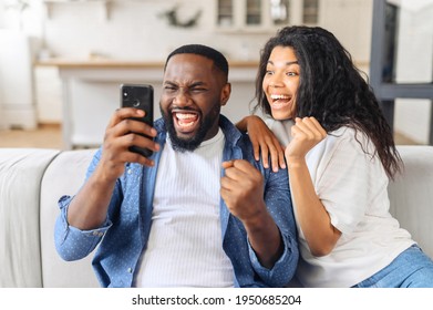 Black Overjoyed Couple Resting On Couch Holding Smart Phone, Celebrating On-line Lottery Win, Bid Betting Victory Moment, Unbelievable Opportunity Or Invitation, Internet Sale, Getting Prize Concept