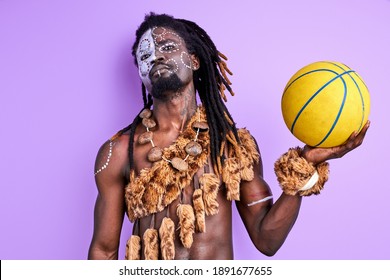 Black Pagan Male With National Make-up Holding Basketball Ball In Hands, Wearing Authentic Traditional Cape On Naked Body. Purple Background