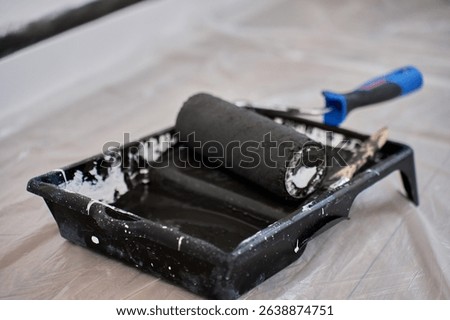 Black paint roller lying in tray filled with black paint on protective film during interior painting process. Preparation for wall painting during home renovation