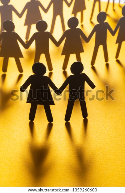Black paper figure of a couple in\
front of a crowd of paper people holding hands on yellow surface.\
Social movement, protest, leadership\
concept.