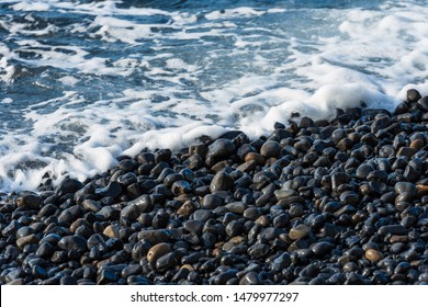 Black Pebbles And The Deep Blue Ocean Closeup At Mavra Volia Beach On Chios, North Aegean Islands, Greece.