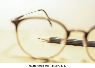 Black Pencil Is On The Brown Notbook In Frame Of Blurred Glasses.