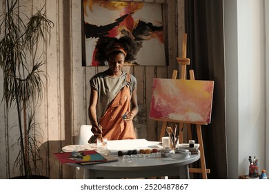 Black person artist, surrounded by paintings, working in cozy home studio with plants and wooden walls. Painting on easel shows a vibrant, abstract design - Powered by Shutterstock