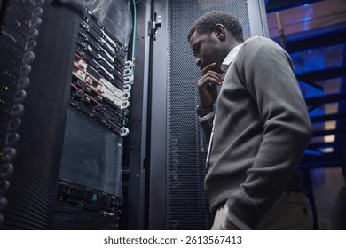 Black person contemplating server maintenance in data center with thoughtful expression examining racks of servers and equipment