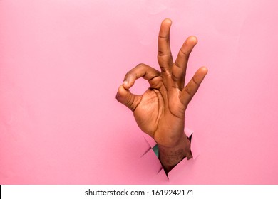 Black Person Hand Through A Hole In A Cardboard Making Ok Sign On Pink Background