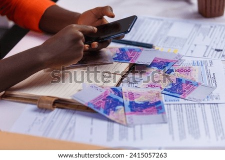 Black person in orange long sleeves shirt working on desk, touching phone screen with notebook and West African CFA franc notes on desk. accounting concept