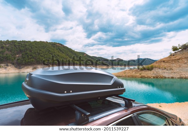 Black plastic car rooftop cargo box or\
roof carrier for traveling. Removable storage container mounted on\
car roof rack. Beautiful nature\
background.