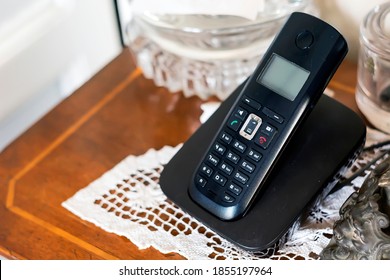 A Black Plastic Cordless Phone Charging On Its Cradle Inside A Classically Furnished Home With Antiques And White Crochet. Outdated Technology