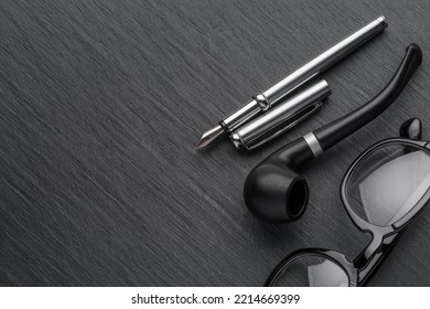 Black plastic eyeglasses with fountain pen and smoking pipe on dark slate background. Copy space - Powered by Shutterstock