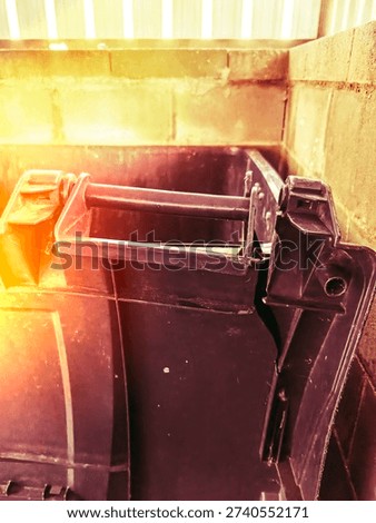 A black plastic garbage bin shows a broken handle and cracks on one side. The bin is placed against a wall in a storage area. The setting is bright and shows sunlight.