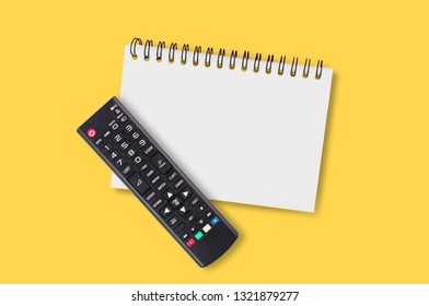 Black Plastic Remote Control For Tv Near Blank Paper Notepad With Spiral Binder Lies On Yellow Table In Home Or Office. Top View.Concept Of Communication Or Multimedia. Copy Space For Your Text