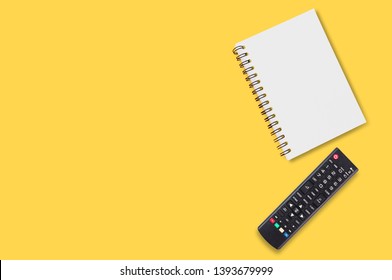 Black Plastic Remote Control For Tv Near Blank Paper Notepad With Spiral Binder Lies On Yellow Table In Home Or Office. Top View.Concept Of Communication Or Multimedia. Copy Space For Your Text