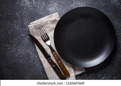 Black Plate, Cutlery And Napkin On Stone Table Top View. Table Setting.