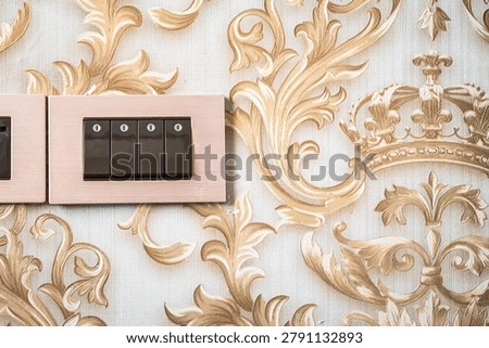 A black platinum light switch is mounted on the wall of the hotel room with a golden, royal crown-patterned wallpaper as a background.