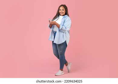 Black Plus Size Woman Using Smartphone Standing On Pink Studio Background, Smiling To Camera. Cheerful Female Using New Mobile Application And Texting. Technology And Gadgets. Full Length
