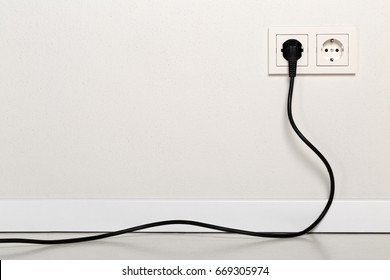 Black power cord cable plugged into european wall outlet on white plaster wall with copy space