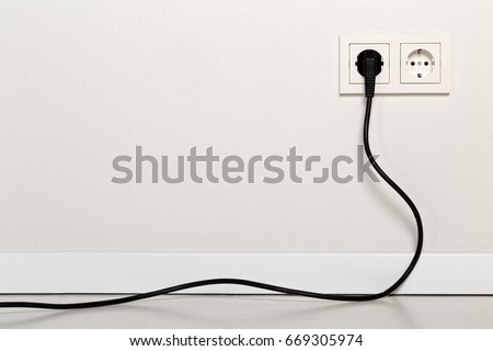 Black power cord cable plugged into european wall outlet on white plaster wall with copy space