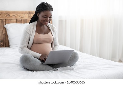 Black pregnant lady using laptop at home, working remotely, browsing internet or shopping online, expectant african woman sitting with computer on comfortable bed, typing on keyboard, copy space