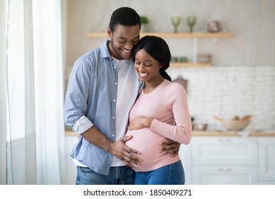 Black pregnant woman and her handsome husband embracing and smiling while spending time together near window at home. Romantic African American couple expecting new baby indoors. Parenthood concept