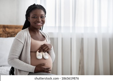 Black Pregnant Woman Preparing For Motherhood, Holding Small Baby Shoes Near Belly, Happy Awaiting African American Lady Sitting On Bed And Smiling At Camera, Enjoying Maternity, Copy Space