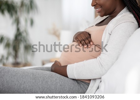 Black Pregnant Woman Sitting On Bed At Embracing Belly, Enjoying Maternity Time, Smiling African Expectant Lady Relaxing In Bedroom At Home, Tenderly Touching Tummy, Side View With Free Space, Closeup