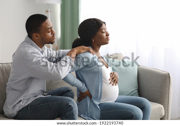 Black pregnant woman suffering from back pain at\
home, attentive spouse comforting her. African expecting lady\
having childbirth labor while sitting with husband on sofa at home,\
copy space