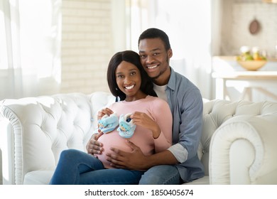 Black Pregnant Woman With Tiny Baby Boties And Her Happy Husband Hugging On Sofa At Home. Future Mom And Dad With Little Shoes Expecting Baby, Embracing On Couch Indoors