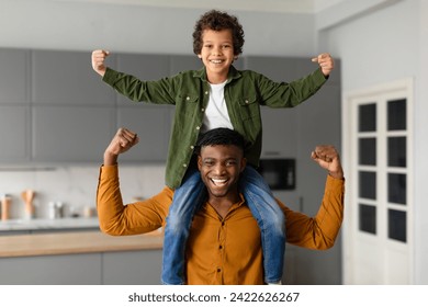 Black preteen boy on his dad's shoulders triumphantly raises his fists in the air, as they both flex their biceps, displaying strength and bonding at home - Powered by Shutterstock