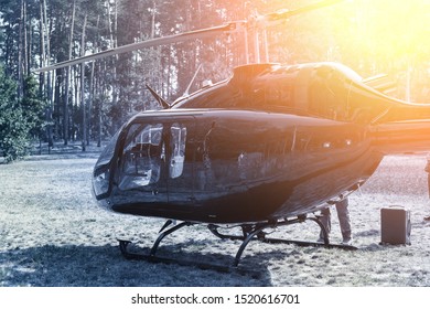 Black Private Modern Luxury Helicopter Standing On Grass Field Near Forest At Country Rural Landscape. Rich Buiness Lifestyle Travel. VIP Aero Taxi Service