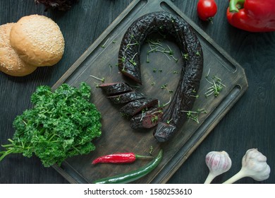 Black Pudding On A Dark Cutting Board Decorated With Greens And Vegetables. Blood Sausage. Home-made Sausage. View From Above. Dark Wooden Background.