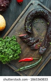 Black Pudding On A Dark Cutting Board Decorated With Greens And Vegetables. Blood Sausage. Home-made Sausage. View From Above. Dark Wooden Background.