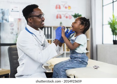 Black qualified pediatrician in white lab coat and eyeglasses examining throat of cute child at modern cabinet. African girl visiting hospital for regular checkup. - Powered by Shutterstock