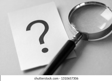Black question mark printed on white paper card on the table next to magnifying glass, viewed in close-up. Search and questioning concept - Shutterstock ID 1310657306
