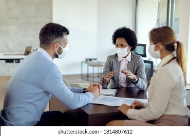 Black Real Estate Agent And A Couple Wearing Protective Face Masks While Communicating During The Meeting In The Office. 