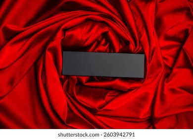 Black rectangular box on luxurious red silk fabric, styled with elegant folds, suitable for product display or advertising mockups.