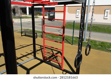 A black and red metal workout structure featuring a vertical ladder and hanging gymnastic rings stands on an outdoor playground with a rubber surface.