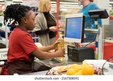 Black Saleswoman Checking Price Of Sunflower Oil While Holding Plastic Bottle Over Cashier Counter On Background Of Two Mature Buyers