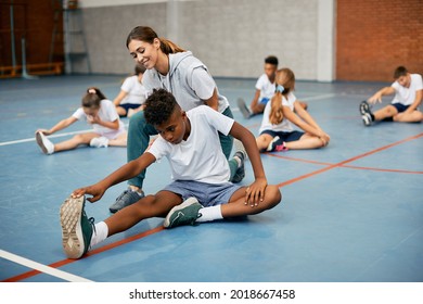 Black School Boy Stretching On The Floor With Help Of PE Teacher While Warming Up Exercise Class. 