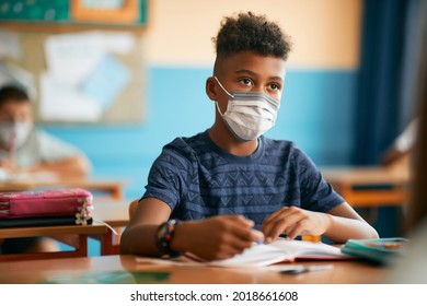 Black schoolboy writing in notebook during a class and wearing face mask due to COVID-19 pandemic.