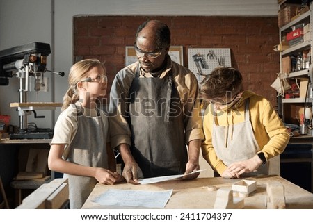 Black senior carpenter teaching two kids woodworking in small workshop and showing them plans