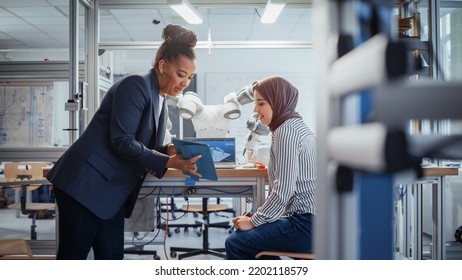 Black Senior Engineer Discussing Robotic Arm With Promising Arabic Female Computer Scientist While Manipulating It With Laptop And Tablet. Manufacturing Facility That Creates High Tech Products.