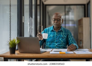 A black senior man in casual clothes is on a video call while sipping coffee at the table. - Powered by Shutterstock