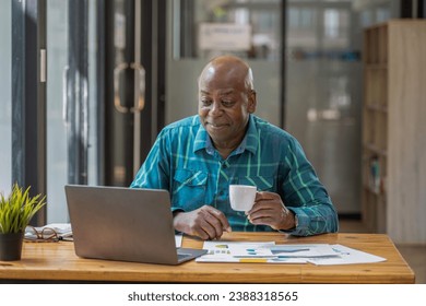 A black senior man in casual clothes is on a video call while sipping coffee at the table. - Powered by Shutterstock