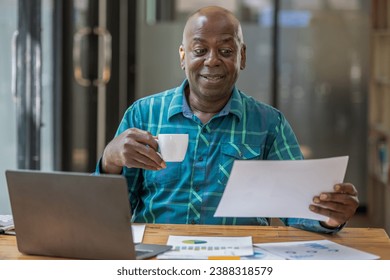 A black senior man in casual clothes is on a video call while sipping coffee at the table. - Powered by Shutterstock