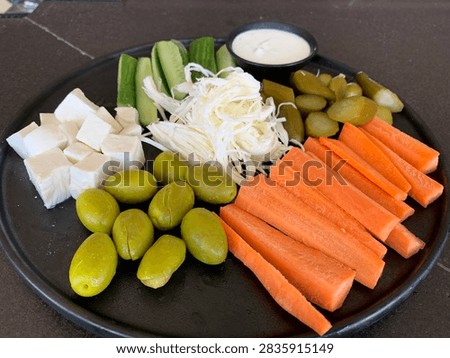A black serving platter arranged with assorted fresh vegetables, cheese cubes, olives, and dip.
A high-angle, close-up photograph of a dark, round platter containing cubed white cheese, large green olives, carrot sticks, sliced cucumbers, shredded white cheese or cabbage, chopped pickles, and a small black bowl of white dipping sauce in the center.