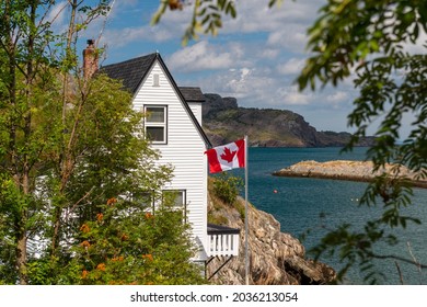 A black shingled roof on the corner of a white wooden cottage. The building is on the edge of a cliff. There's a Canadian flag flying on the patio overlooking the ocean with mountains. 