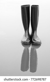 Black Shiny Rubber Boots Stand In A Puddle Of Rain And Reflect In The Water. Proper Footwear For Wet Weather. A Black And White Photography. Rubber Boots Become Art.
