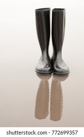 Black Shiny Rubber Boots Stand In A Puddle Of Rain And Reflect In The Water. Proper Footwear For Wet Weather. Rubber Boots Become Art.
