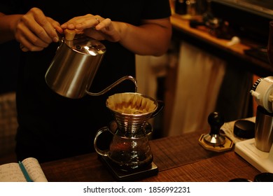 Black Shirt Barista Pouring Hot Water On Coffee Ground With Filter From Silver Teapot To Transparent Chrome Drip Maker On Wooden Table Cafe Shop. Drip Brewing, Filtered Coffee.
